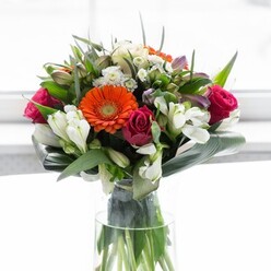 A floral composition of remarkable grace, this bouquet echoes the vibrant yet peaceful atmosphere of Petts Wood. Creamy, soft peach gerbera daisies trace a gentle arc around the centerpiece, their bright orange siblings bursting forth like lanterns beside them, recalling the warmth shared at local gatherings near Station Square. Enclosed by bold, rich magenta roses, the display gains both depth and flourish, while clusters of ivory alstroemeria and sprays of dainty white roses suggest a tranquil, garden-fresh ambiance. Sprinkles of pink filler flowers and loose, airy whites add delicacy and volume, like the wild blooms lining woodland paths. Encasing it all, a cascade of deep green foliage gives structure, with paddle-shaped aspidistra leaves expertly curved as a lush collar at the base-echoing the protective embrace of Petts Wood's famous trees. The arrangement stands proud in a clear glass vase, where vibrant green stems are visible, bathed in gentle light that lifts every hue and underscores the local florist's fine attention to detail. From the bouquet drifts a subtle scent-a harmonious blend of roses, lilies, and fresh greenery. Ideal for celebrations, reunions, or sending messages of encouragement, this arrangement from Petts Wood Florist brings together skill, community spirit, and optimism in every carefully arranged petal.